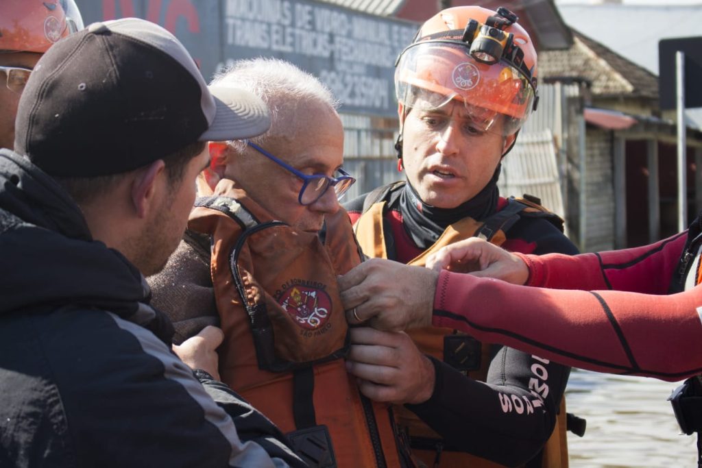 bombeiro fardado, com capacete, auxilia idoso em uma operação de resgate. Ambos usam colete salva-vidas