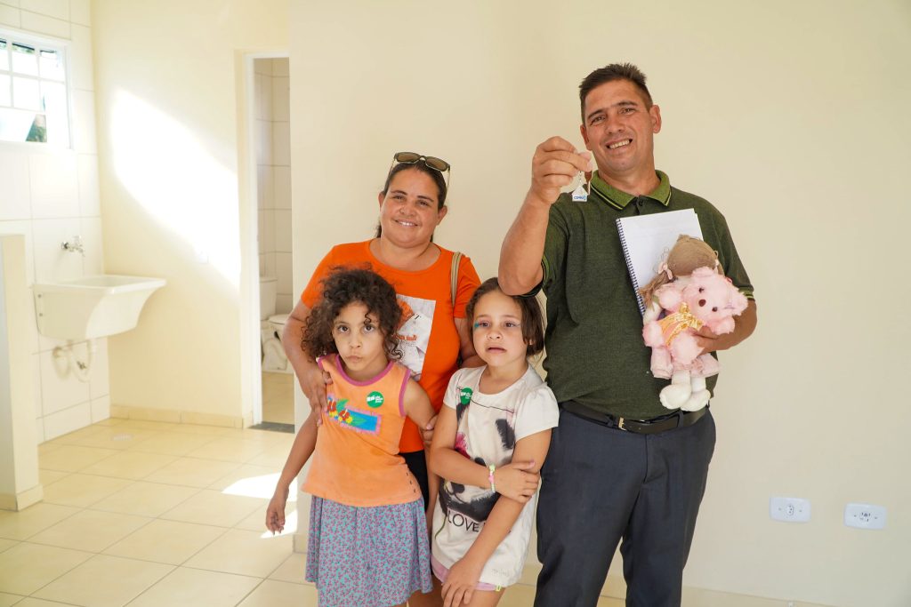 família, com um pai, uma mãe e duas crianças meninas com sorriso no rosto mostranda a chave de sua nova moradia