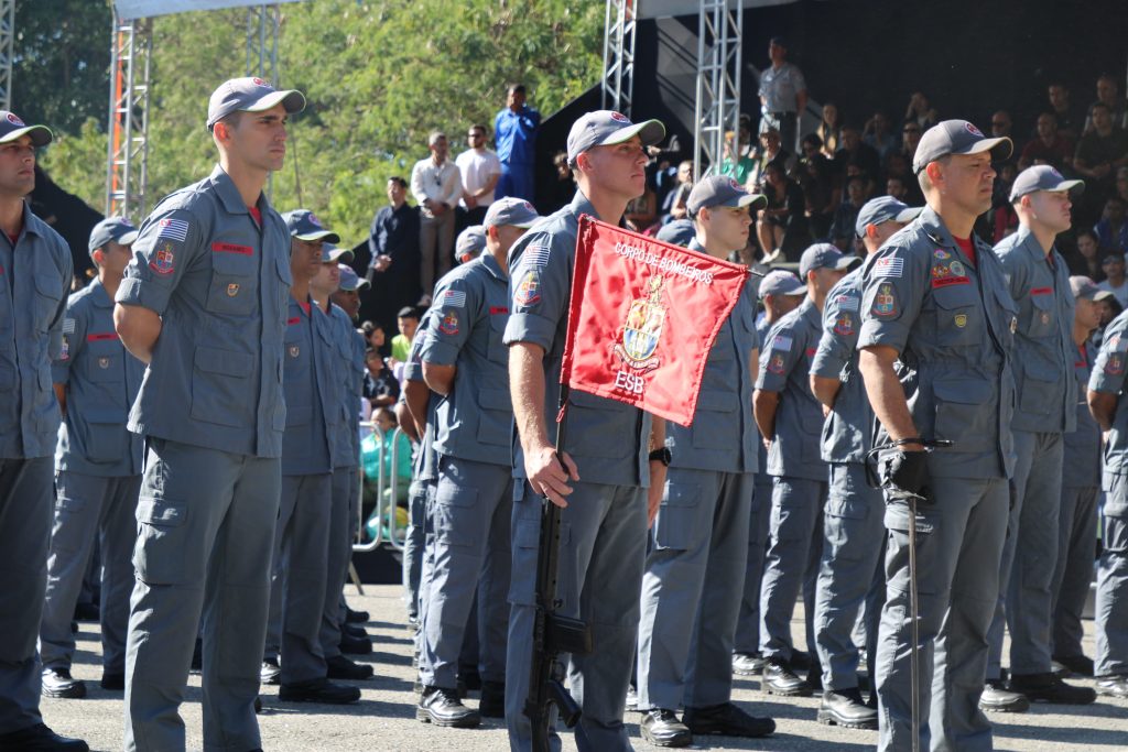 integrantes do Corpo de Bombeiros de São Paulo fardados e enfileirados. Um deles segura uma bandeira vermelha da corporação