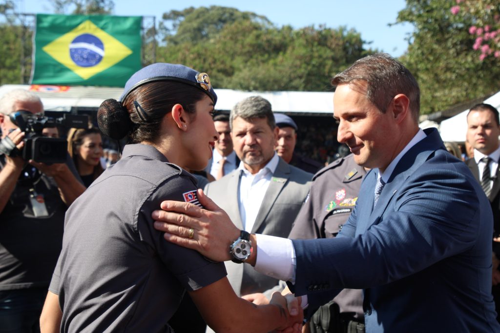 fotografia do secretário de segurança pública de São Paulo, Guilherme Derrite, um homem branco, de terno e cableo curto apertando a mão de uma policial militar fardada